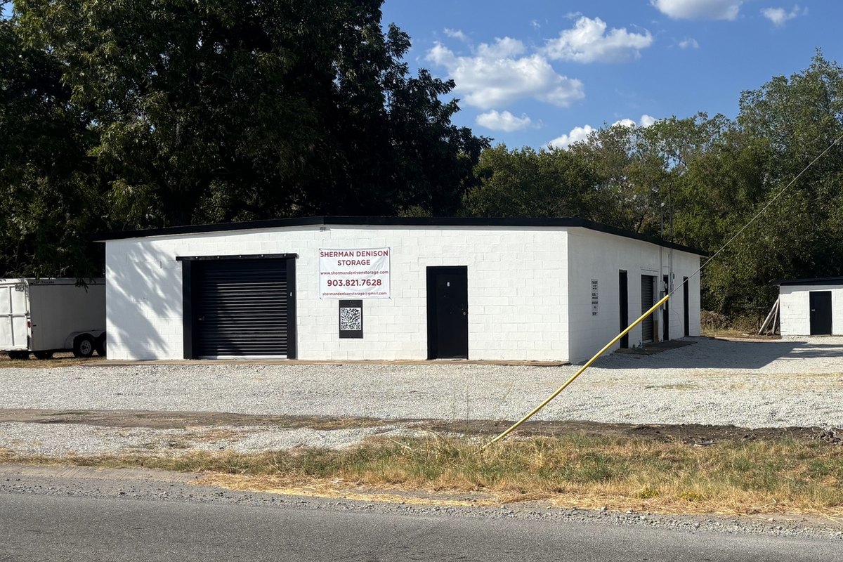 White cinder-block storage building with a black roll-up door and a banner, seen straight on from across the road
