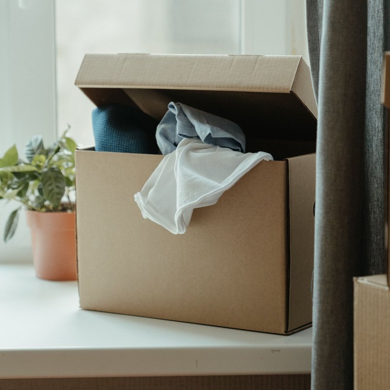 Open cardboard box of folded clothes and stacked boxes by a window with a houseplant