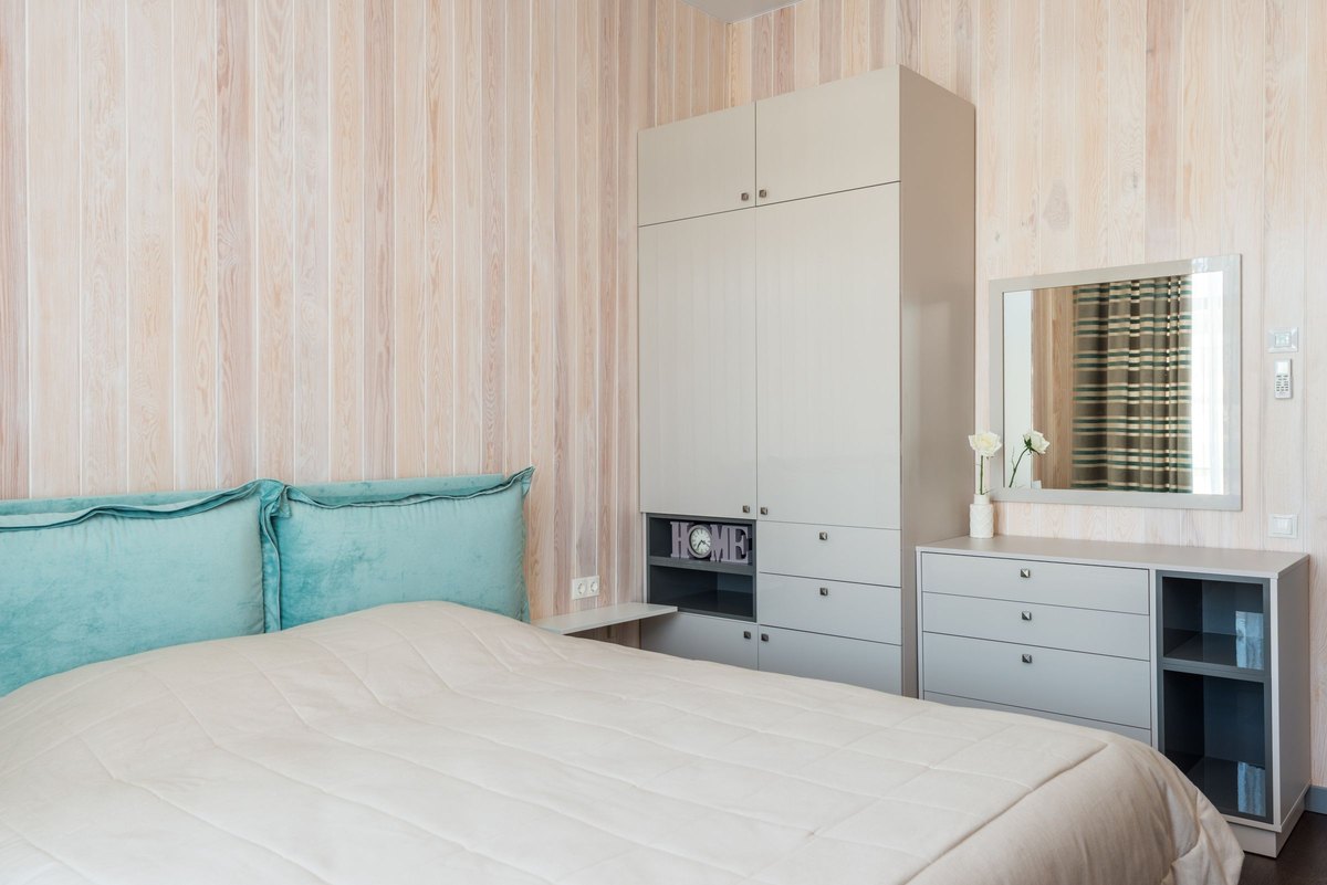 Made bed with teal velvet headboard pillows beside a gray wardrobe and dresser in a bright wood-panelled bedroom