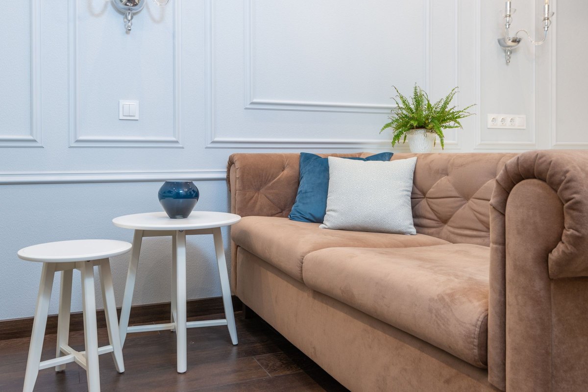 Tan tufted velvet sofa with blue and gray pillows beside two white side tables and a fern in a bright room
