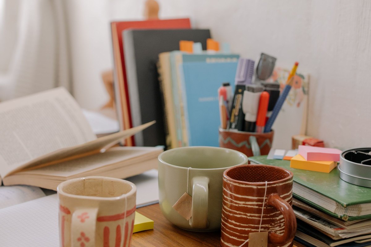 Ceramic mugs, an open book, notebooks and textbooks on a wooden study desk by a window