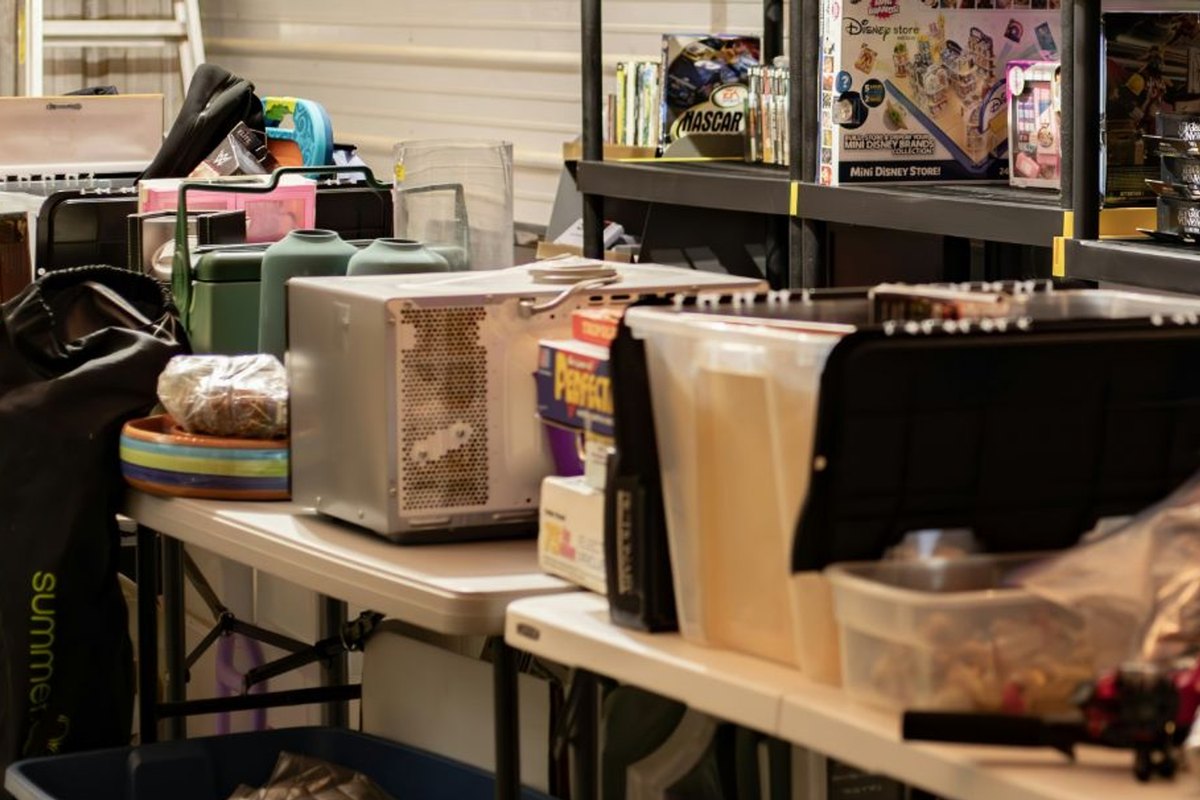 Folding tables with a microwave, clear plastic tubs and a black case inside a crowded storage unit