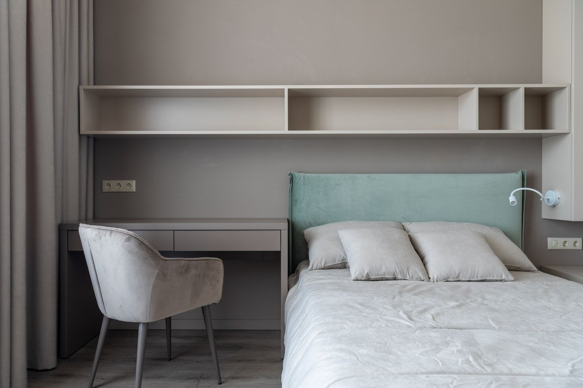 Made bed with a mint headboard beside a built-in desk and a gray armchair in a tidy bedroom