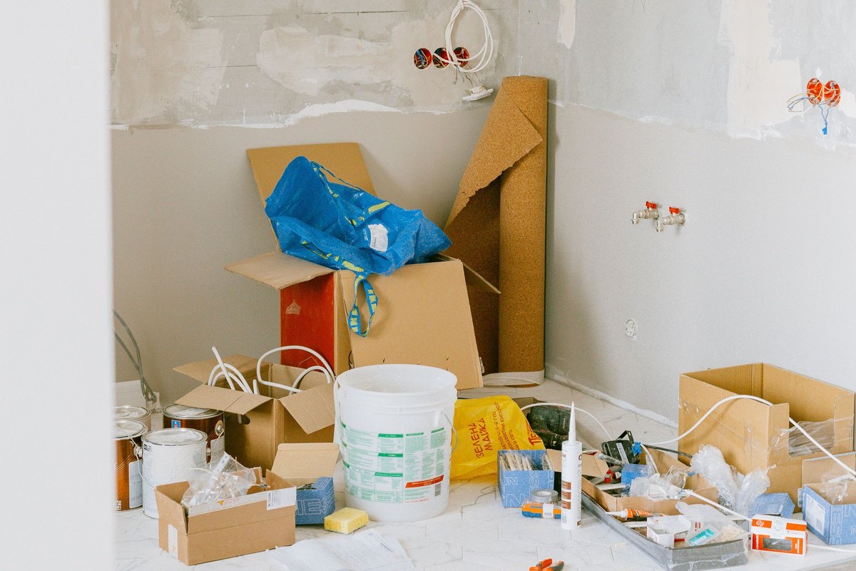 Cardboard boxes, a bucket and paint cans on the floor of a room under renovation with bare plaster walls