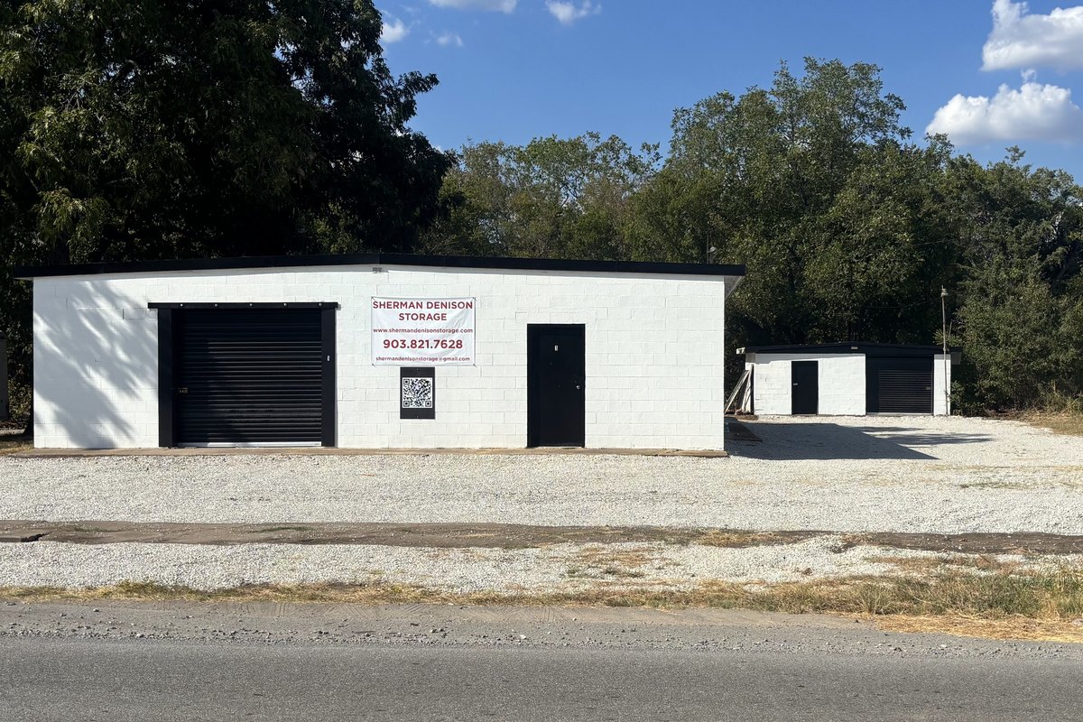 Straight-on view of the white storage building with its black roll-up door and a small shed beside it on a white gravel lot