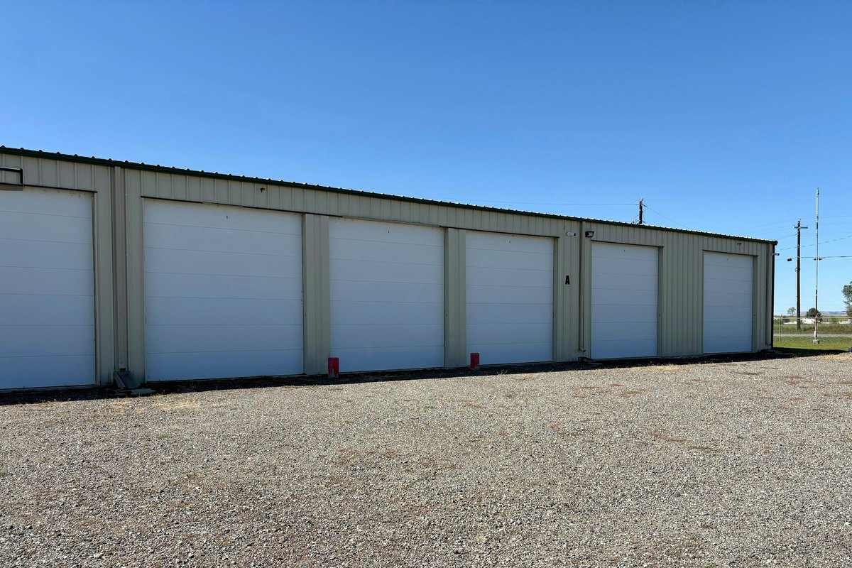 Beige metal storage building with six white garage-style doors on a gravel lot under a blue sky